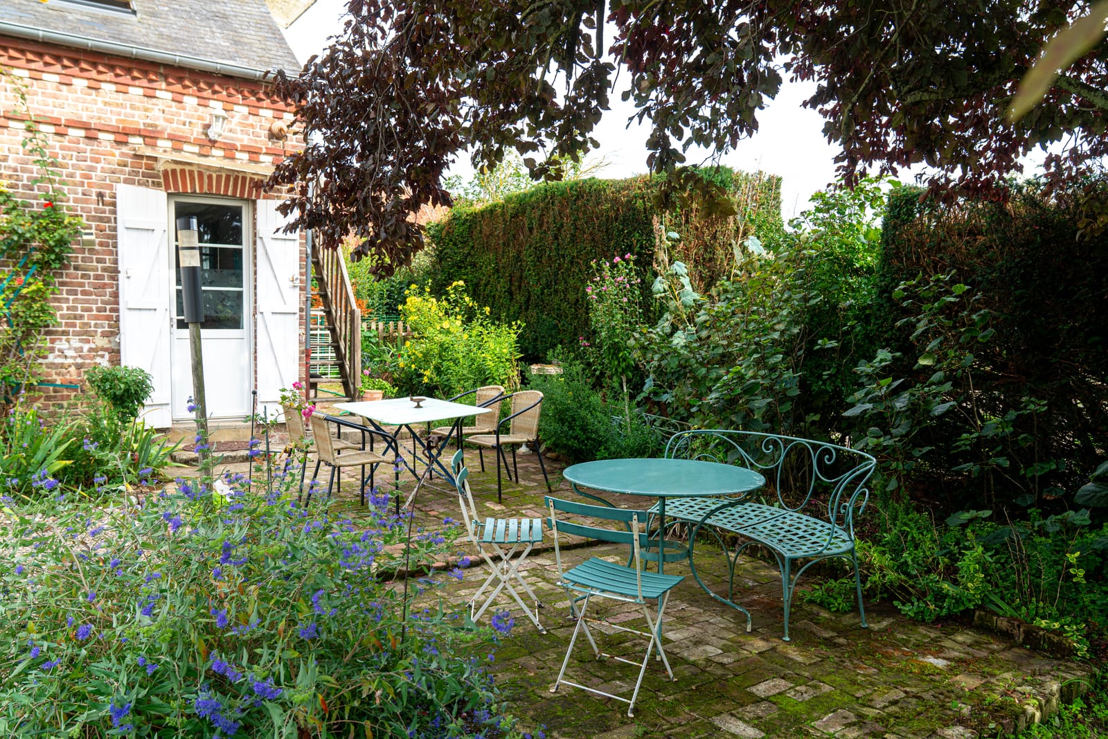Le jardin de la maison : tables et chaises en fer, banc courbe et fleurs bleues, devant la porte aux volets blancs